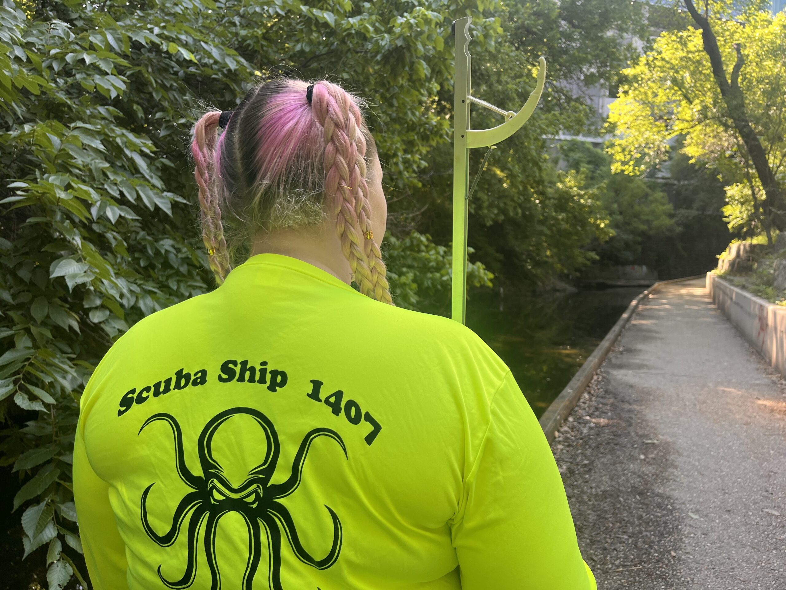 Person wearing a bright yellow shirt with 'Scuba Ship 1407' and an octopus design on the back, standing beside a pathway near a creek surrounded by greenery.